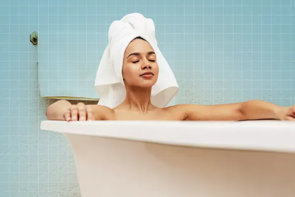 डोक्यावर पांढरा टॉवेल घातलेली स्त्री डोळे मिटून आंघोळ करताना आराम करते